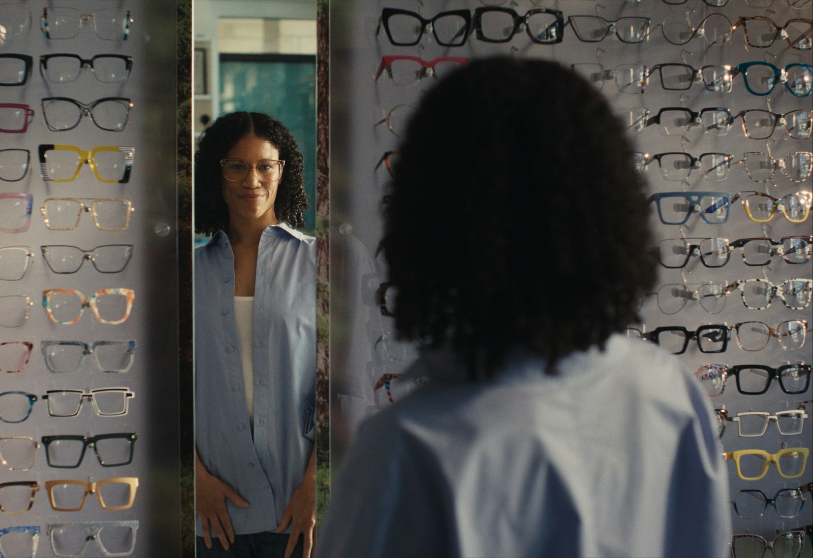 Smiling woman trying on new eyeglasses, seen in the mirror at an optical shop with a wall of frames